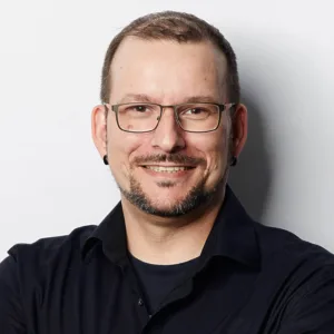 Ein Porträt eines lächelnden Mannes mit Brille, kurzem, dunklem Haar und einem kurzen Bart. Er trägt ein schwarzes Hemd und hat schwarze Ohrstecker. Der Hintergrund ist weiß.